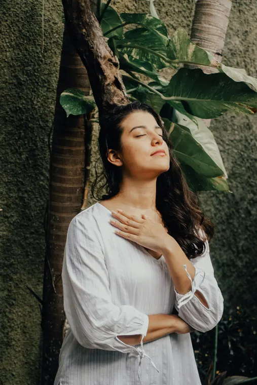 A woman in a white dress leaning against a tree, eyes closed, a hand resting on her chest